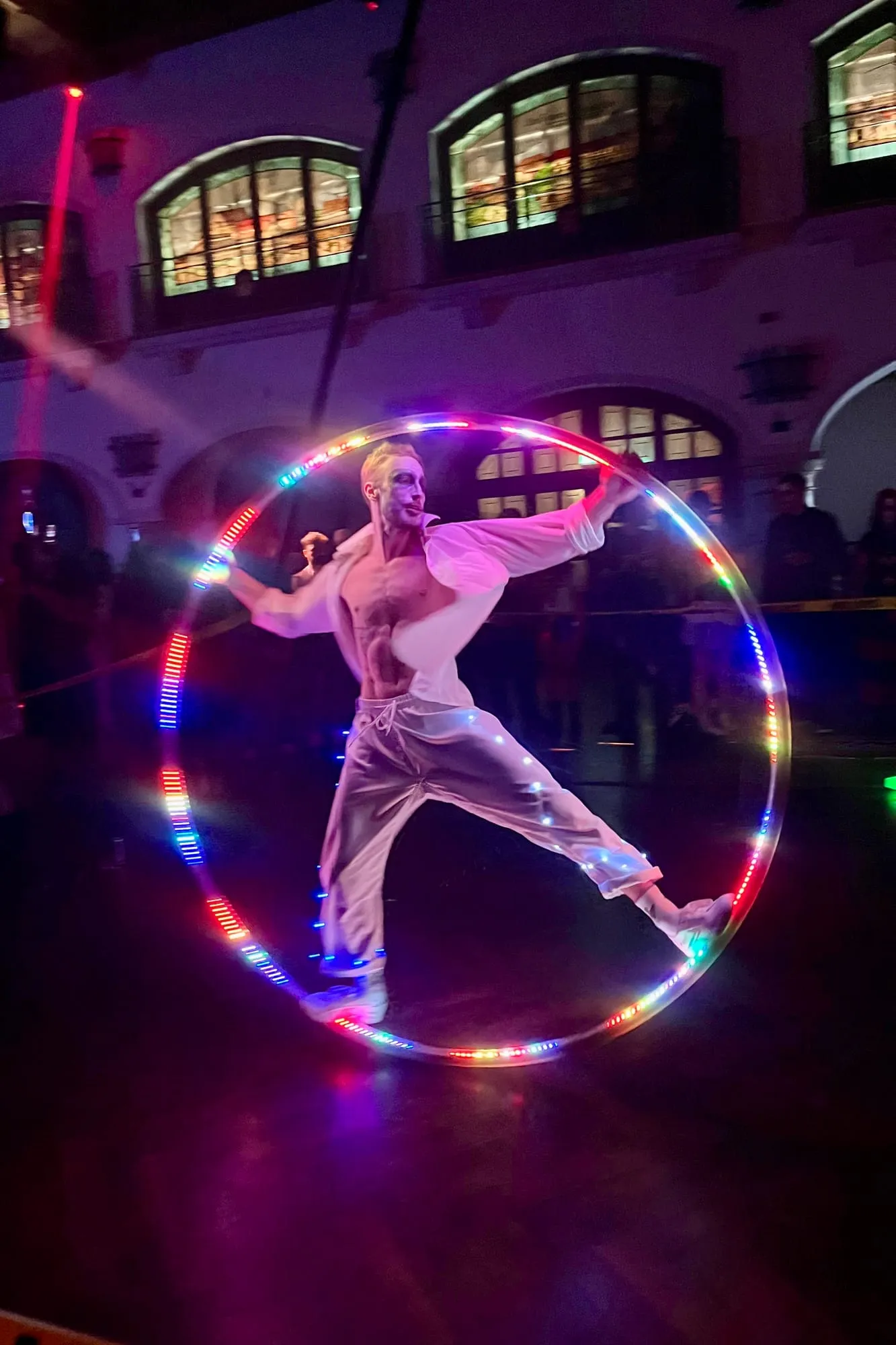 Im Cyr Wheel mit bunten LED-Lichtern, Arme und Beine weit gestreckt, in einer dunklen Halle