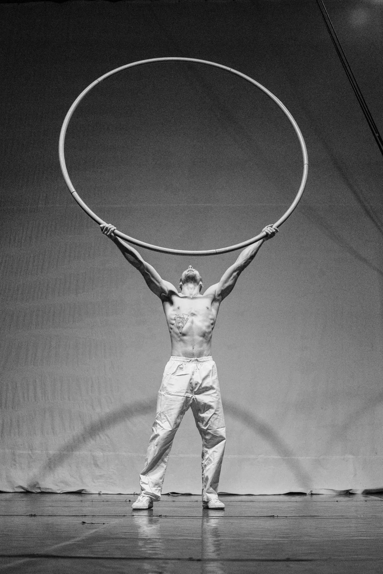 Schwarzweiß: mit nacktem Oberkörper auf der Bühne, das Cyr Wheel mit beiden Armen hoch über den Kopf gestemmt